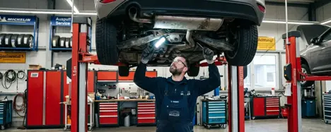 Mechaniker inspiziert Unterboden eines silbergrauen Gebrauchtwagens auf einer Hebebühne mit LED-Taschenlampe